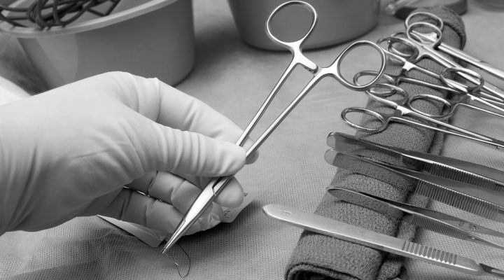 Technician picks up needle holder and suture from sterile field in operating room.